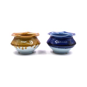 Pode incluir: Dois vasos de cerâmica, um com um esmalte marrom e o outro com um esmalte azul. Ambos os vasos têm uma borda festoneada e um interior branco.