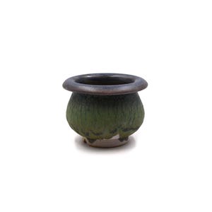May include: A small, rounded ceramic pot featuring a wide, dark brown rim that transitions into a textured, moss-green body. The base of the pot is unglazed, revealing a light beige ceramic material with dark, irregular markings along the bottom edge.
