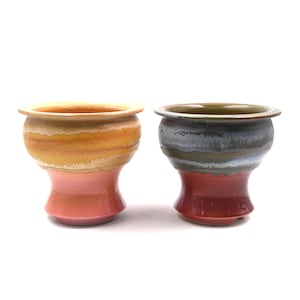 May include: Two ceramic vessels with wide rims and flared bases. The left vessel features horizontal bands of yellow, orange, and pink glaze. The right vessel displays a dark green and grey mottled upper section with a solid red base glaze.