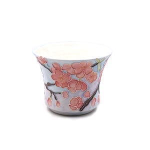May include: A ceramic planter with a light blue background and a floral design. The design features pink cherry blossoms and dark brown branches. The planter has a wide opening and a slightly flared shape.