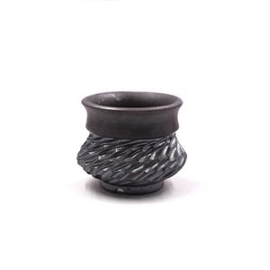Pode incluir: Uma caneca de cerâmica cinza escuro com um padrão texturizado, semelhante a um tecido, na metade inferior. A parte superior é lisa e cilíndrica. A caneca é projetada com uma borda larga e arredondada.