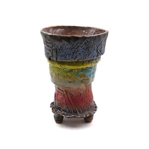 Könnte beinhalten: Eine handgefertigte Keramikvase mit einem strukturierten, geschichteten Design. Die Vase hat einen dunkelblauen oberen Teil, der in Grün-, Gelb- und Rottönen übergeht. Sie steht auf drei kleinen Füßen und hat eine breite Öffnung, ideal für Blumen.