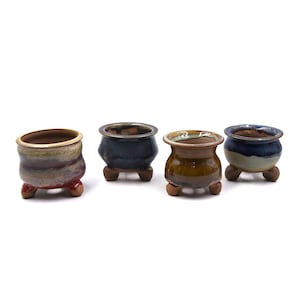 May include: Four small, ceramic bonsai pots in a row. Each pot has a unique color scheme, including shades of blue, brown, and red. The pots are round with a flared rim and sit on three small feet. The pots are ideal for small plants.