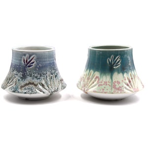 Könnte beinhalten: Zwei Keramikbecher mit einem ausgestellten Boden und einem strukturierten Design. Der Becher links ist blau und weiß mit einem violetten Blattdesign. Der Becher rechts ist grün und weiß mit einem rosa und weißen gesprenkelten Design.