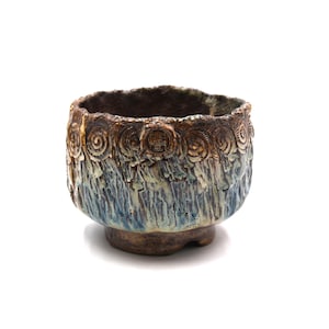 May include: A handcrafted ceramic bowl with a textured surface and a unique design. The bowl features a brown rim with spiral and embossed details, transitioning to a blue and cream-colored body. The base is a darker brown.