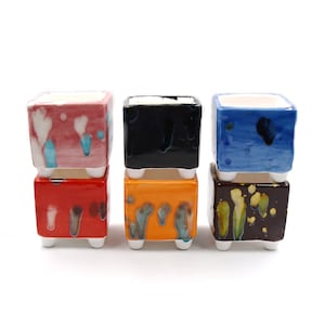 May include: Six small square ceramic planters in various colors, stacked in two rows of three. The planters have a unique, abstract glaze pattern in shades of red, orange, yellow, blue, black, and white.