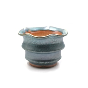 May include: A small, handcrafted ceramic planter with a wavy rim. The exterior features a gradient of blue-green hues, with a darker band around the middle. The interior is a warm orange color. The planter is designed for small plants.