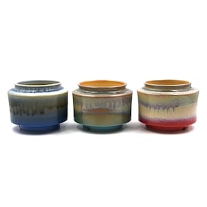 Puede incluir: Tres macetas de cerámica con forma cilíndrica y una abertura ancha. Las macetas tienen una combinación de colores de dos tonos, con un color base y un esmalte que gotea por los lados. Los colores incluyen azul, verde azulado y rojo. Las macetas están sobre un fondo blanco.