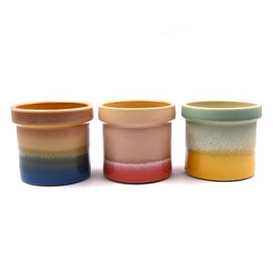 May include: Three ceramic pots with a cylindrical shape and a wide rim. Each pot features a gradient color scheme, with hues of blue, orange, pink, and yellow. The pots are arranged side by side against a white background.