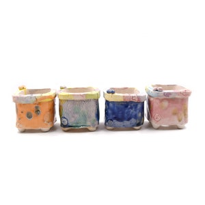 May include: Four small, square ceramic planters with different colourful patterns and glazes. The planters have a slightly raised rim and three small feet on the bottom.