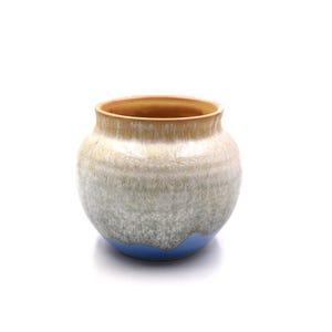 May include: A handcrafted ceramic vase with a rounded shape. The vase features a gradient of colors, starting with a tan rim, transitioning to a white and gray body, and finishing with a blue base. The pottery has a textured appearance.