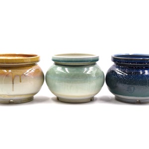 May include: Three ceramic pots in different colors and glazes. The pot on the left is a light brown with a yellow rim. The middle pot is a light green with a blue rim. The pot on the right is a dark blue with a white speckled glaze.
