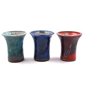 May include: Three handmade ceramic vases in teal, blue, and red. Each vase has a flared rim and a textured surface. The vases are displayed against a white background. These decorative vases are suitable for home decor.