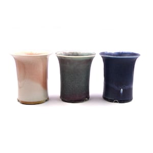 Puede incluir: Tres macetas de cerámica en diferentes colores. La maceta de la izquierda es de color rosa claro y blanco. La maceta del centro es de color verde oscuro y morado. La maceta de la derecha es de color azul oscuro.