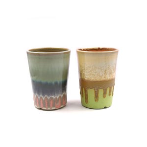 May include: Two ceramic mugs with a drip glaze design. The mug on the left has a green, blue, and pink glaze. The mug on the right has a yellow, brown, and green glaze.