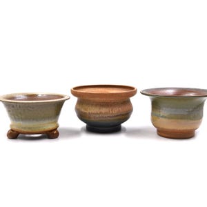 May include: Three ceramic plant pots with a rustic, glazed finish. The pots vary in shape and size, with earthy tones of brown, green, and yellow. One pot has small feet. The pots are suitable for small plants and succulents.