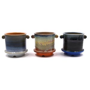 May include: Three cylindrical ceramic planters with small spiral handles on each side. The left planter has a blue and grey gradient, the center one features a multi-colored drip glaze, and the right one is dark black with a bright blue base.