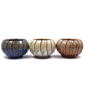 May include: Three small, round ceramic bowls with a ribbed, fluted exterior texture. The left bowl is dark blue and grey, the center bowl is white with brown crackle details, and the right bowl is a mix of green, brown, and orange tones.