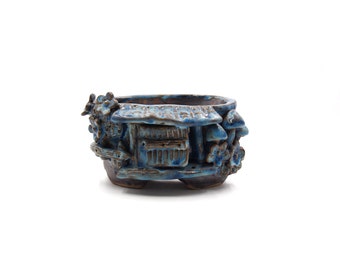 May include: A blue and brown ceramic planter with a whimsical design featuring a small house, mushrooms, and flowers. The planter has a rounded shape and a textured surface.