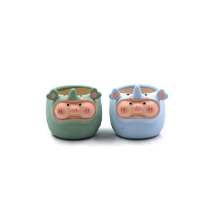 May include: Two ceramic planters, one in light blue and the other in green, each shaped like a rhino. The planters have a pig-like snout and ears, with a small horn. The planters are suitable for small plants or succulents.