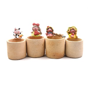 Peut inclure: Quatre petits pots cylindriques en céramique brun clair, chacun surmonté d'une figurine miniature différente. Les figurines comprennent une vache, une fille avec un nœud, une fille avec une fraise et une fille avec une fleur.
