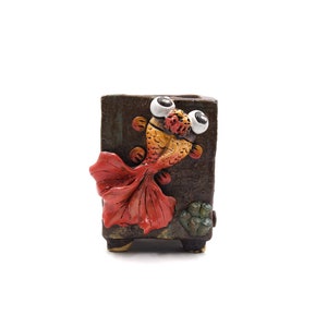 May include: A whimsical, rectangular ceramic planter featuring a detailed, three-dimensional goldfish design. The fish has large, googly eyes, orange and yellow scales, and a vibrant red tail. The planter is brown with a textured finish.