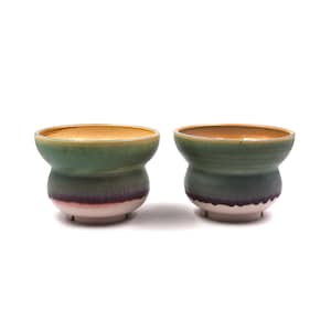 以下が含まれることがあります： 広い縁と丸みを帯びた胴体を持つ2つの陶器製カップ。内側は温かみのある茶色で、外側は緑、濃い紫、そして底部のオフホワイトへと変化するグラデーションが施されています。それぞれのカップには小さな円形の高台が付いています。