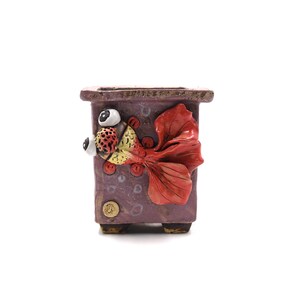 May include: A purple ceramic planter with a 3D red, yellow, and white fish design. The fish has large eyes and a whimsical expression.