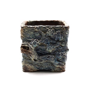 May include: A square ceramic planter with a textured, bark-like exterior. The planter is primarily blue with brown and grey accents, and features a relief design of flowering branches. The interior is a darker brown.