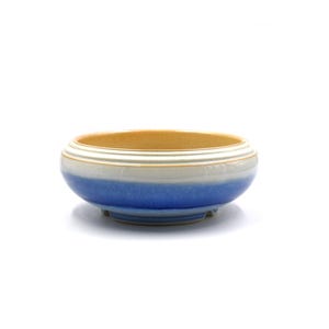 Puede incluir: Un cuenco de cerámica pequeño y redondo con un diseño en capas. El cuenco presenta un interior de color beige claro, una banda blanca y un degradado de tonos azules en el exterior. El cuenco tiene un perfil bajo y un diseño simple y elegante.