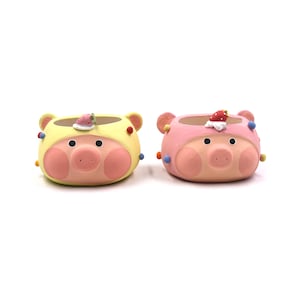 May include: Two oval-shaped planters shaped like pig faces, one in yellow and one in pink. Each planter features a small strawberry decoration on top, round black eyes, pink cheeks, and small colourful dots on the sides of the pig-shaped exterior.