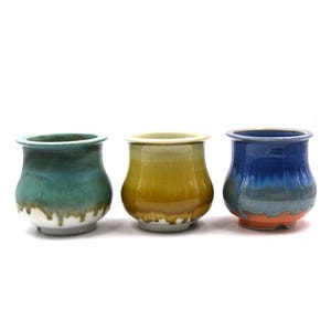 May include: Three small ceramic cups with a rounded, bulbous shape and a flared rim. The first cup features a teal and white drip glaze, the second a yellow and brown gradient, and the third a deep blue and orange drip glaze finish.