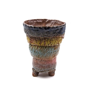 May include: A handmade ceramic vase with a distinctive layered design. The vase has a textured finish and is painted in various colours, including brown, blue, yellow, and pink. The vase has three small feet and a wide opening.