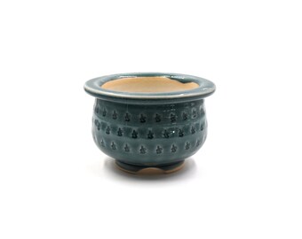 May include: A teal ceramic planter with a textured, repeating floral pattern. The small, round pot has a wide rim and a light beige interior. The base is a darker shade of teal. Ideal for small plants or succulents.