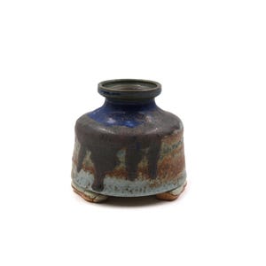 May include: A small, handcrafted ceramic vase with a dark blue top and a brown, dripped glaze effect. The vase has a rounded body and sits on three small feet. The color palette includes shades of blue, brown, and tan.
