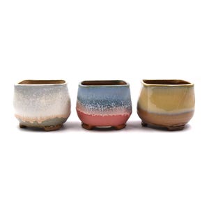 May include: Three ceramic plant pots with square openings and rounded bodies. The pots are in shades of blue, yellow, and white with a speckled glaze. Each pot has three small feet.