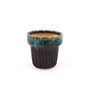 May include: A ceramic flower pot with a dark brown exterior and a teal rim. The rim features a floral pattern. The pot has vertical ridges and the text "T's Garden" is visible on the side. The interior is a light brown color.