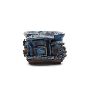 May include: Blue ceramic planter shaped like a small house with a door and a window. The planter has a textured surface and is decorated with small flowers and a bird.