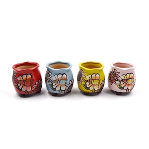 May include: Four small ceramic flower pots with painted floral designs. The pots are red, blue, yellow, and pink. Each pot has three small feet.