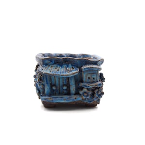 May include: A blue ceramic planter with a decorative design of a small house with a window and a roof. The planter has a textured surface and is decorated with small flowers.