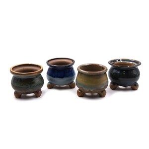 Puede incluir: Cuatro pequeñas macetas de cerámica para bonsái en varios colores: verde, azul, amarillo y negro. Cada maceta tiene forma redondeada, boca ancha y tres patitas. Acabado brillante.