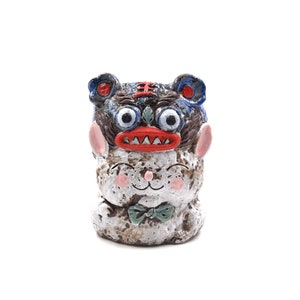 May include: A ceramic figurine of a white cat wearing a blue and red lion mask. The cat has a green bow tie and a white face with pink cheeks. The figurine is sitting on a white background.