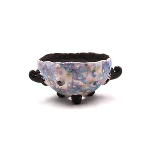 May include: A small, round bowl with a textured, multi-colored exterior featuring shades of light blue, pink, and white. The bowl has a dark, matte black interior, three small black feet, and two decorative, curled black handles on the sides.