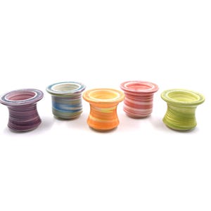 May include: Five colorful, ceramic spool-shaped candle holders. Each holder has a unique swirl pattern in shades of purple, blue, orange, pink, and green. The holders are arranged in a row against a white background.