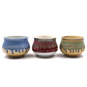 May include: Three ceramic cups with rounded bodies and flared rims. The left cup is blue with a tan base and drip glaze. The center cup features horizontal bands of red, white, and dark gray. The right cup is green with a tan base and drip glaze.