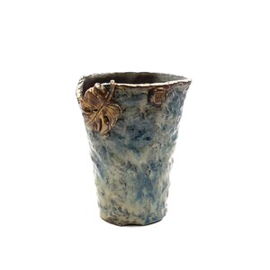 Pode incluir: Um vaso de cerâmica texturizado azul e creme com um detalhe de folha dourada. O vaso tem uma forma áspera e orgânica e um topo ligeiramente alargado. Um pequeno emblema dourado é visível na lateral.