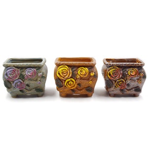 May include: Three ceramic flower pots with different floral designs. The pots are square and have a rustic look. The first pot has a green glaze with purple and pink flowers. The second pot has a brown glaze with yellow flowers. The third pot has a brown glaze with orange flowers.