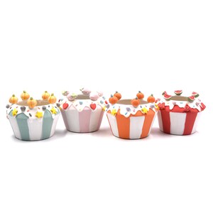 May include: Four ceramic planters shaped like cupcakes with striped bases in light blue, pink, orange, and red. Each features white icing-like rims decorated with small sculpted elements including pumpkins, hearts, strawberries, and watermelon slices.