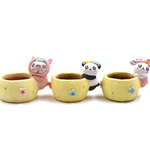 May include: Three yellow ceramic planters with cartoon animal figures. The planters are decorated with small flowers and have a light brown interior. The animals include a pig, a panda, and a bunny.
