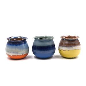 May include: Three small ceramic pots with scalloped rims and rounded bodies. The first pot features a blue, white, and orange gradient. The second pot is a solid blue gradient. The third pot displays a brown, white, and yellow color scheme.